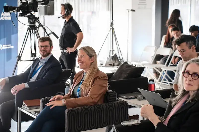 Panelists sit in patterned armchairs, smiling during the AquaHacking Prairies event. A camera operator with a headset stands behind them next to a professional broadcast camera. In the background, audience members sit in rows of white chairs in a bright, modern industrial space.