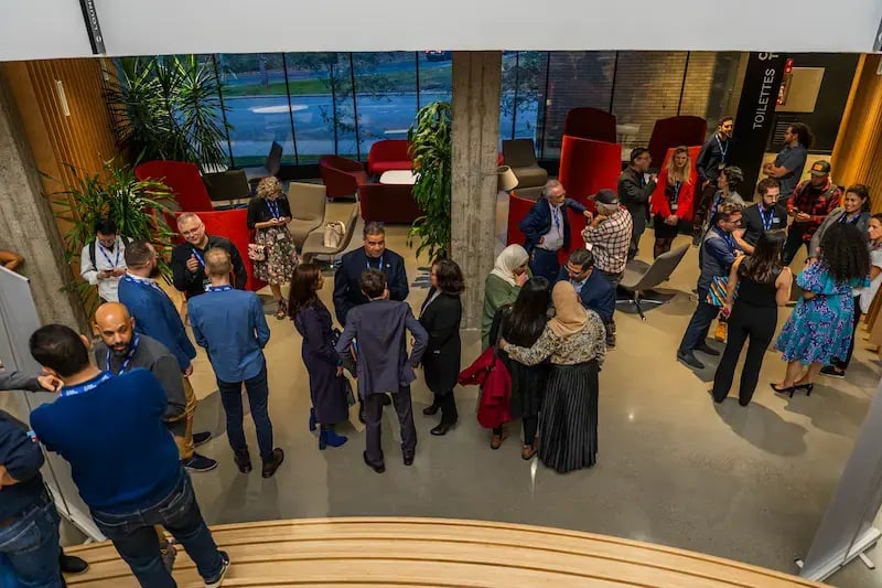 From a high-angle view, participants network in a modern lobby during AquaHacking Québec 2023. Groups of people in professional attire and event lanyards talk near red and grey lounge seating. Large windows in the background look out onto a street at dusk.