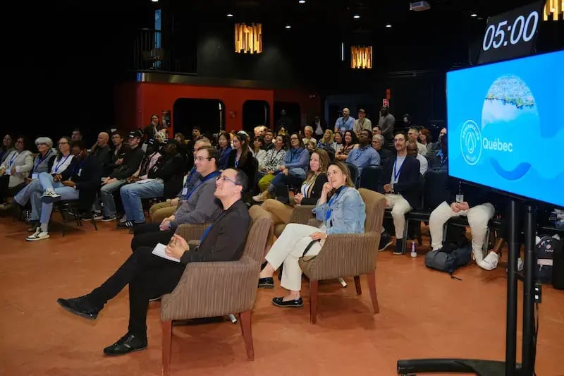 A large audience watches the AquaHacking Québec 2025 finale in a dimly lit theater. In the foreground, judges sit in armchairs facing a digital screen that displays the event logo and a "05:00" countdown timer. The atmosphere is professional and focused.