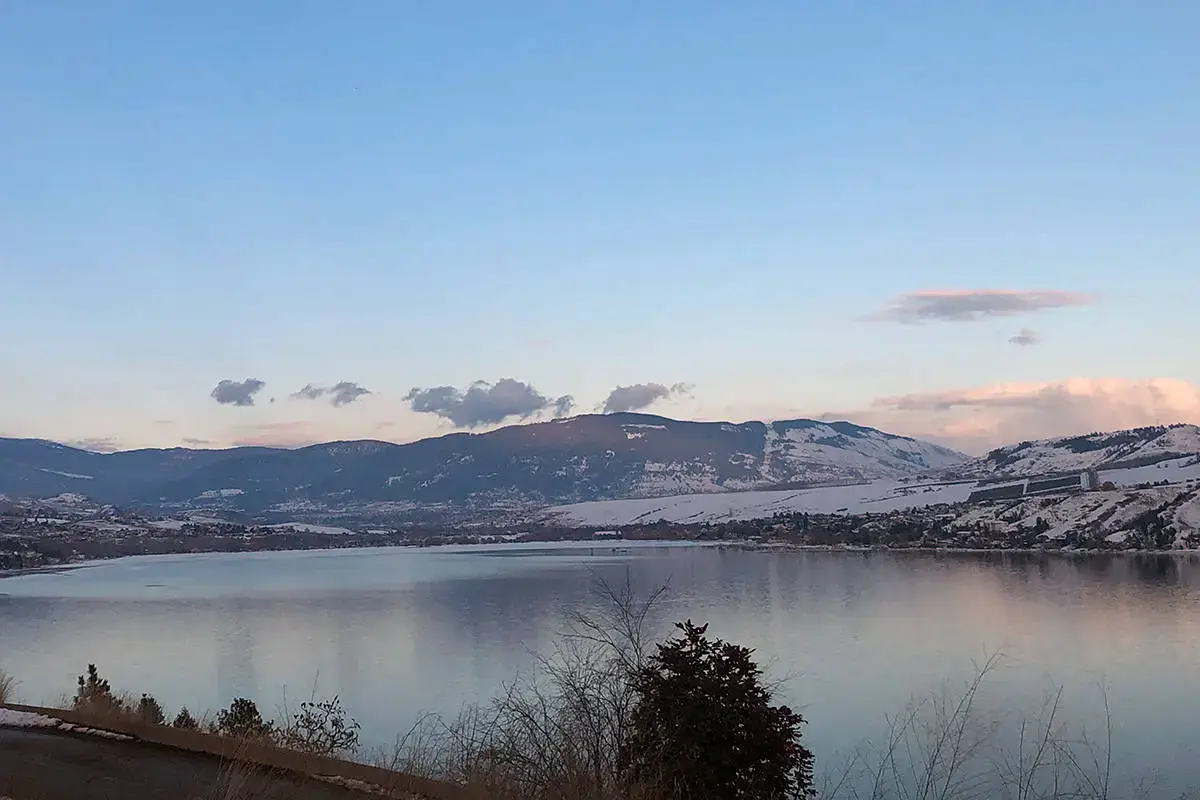 A serene view of Okanagan Lake with snowy mountains in the background, reflecting the calm blue sky and scattered clouds at dusk.