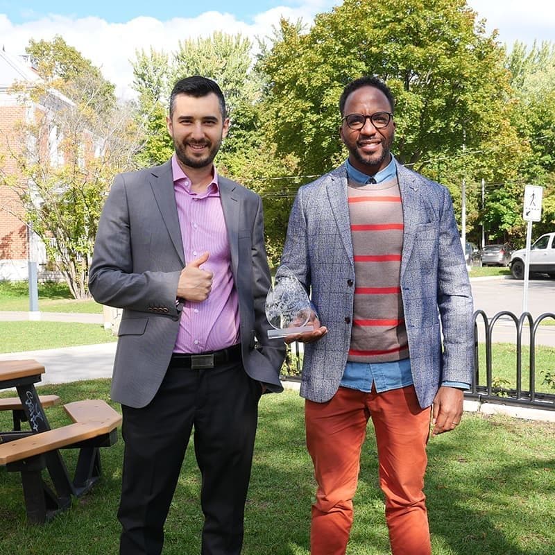 Two men smiling, one holding an award outdoors.