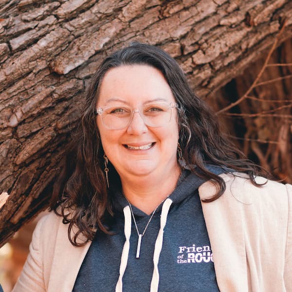 Ashley Flintoff smiles, wearing clear glasses, a dark hoodie printed with Friends of the ROUGE, a light beige jacket, and a bar pendant necklace. She has long, dark wavy hair and poses outdoors in front of a textured tree trunk.