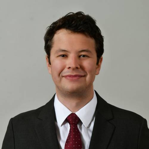 John Schmidt smiles slightly in a studio portrait, wearing a dark grey suit jacket, crisp white collared dress shirt, and a deep red patterned tie. He has dark brown, slightly wavy hair and stands centered against a neutral light grey background.