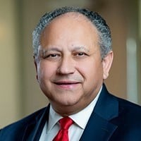 Carlos Del Toro smiles in a formal portrait, wearing a dark blue suit jacket, a white dress shirt, and a bright red tie. He has short grey hair and a warm, approachable expression against a softly blurred, neutral background.