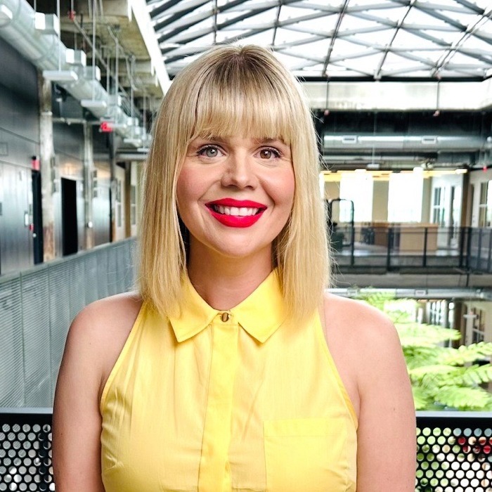 Hilary Doe smiles with bright red lipstick, wearing a sleeveless yellow collared shirt. She has shoulder-length blonde hair with straight bangs. She stands on a balcony inside a modern, industrial-style building featuring a glass atrium roof and exposed ceiling fixtures.