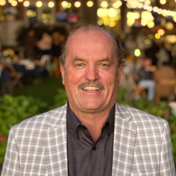 Close-up portrait of Mike Skinner smiling, wearing a dark button-down shirt beneath a light gray plaid blazer. He has a dark mustache and light eyes. Warm string lights create a soft, blurred bokeh background behind him outdoors at night.