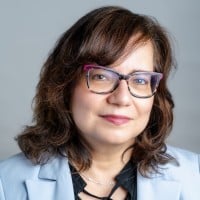 Soula Chronopoulos smiles softly in a close-up portrait. She wears cat-eye glasses with pinkish-purple rims, a light blue blazer, and a black top. Her shoulder-length wavy brown hair is styled with side bangs against a neutral gray background.