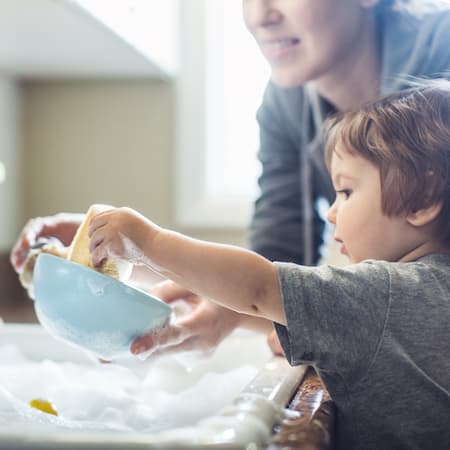 A young child washing a bowl with the help of an adult at a kitchen sink filled with soapy water, illustrating small everyday actions for sustainability and water conservation.