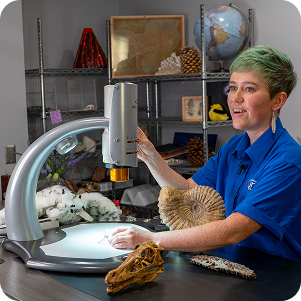 A woman with green hair uses a digital microscope to examine specimens in a science lab. On the desk are an ammonite fossil and a dinosaur skull. The background features shelves with a globe, a volcano model, and various geological samples. A woman with green hair uses a digital microscope to examine specimens in a science lab. On the desk are an ammonite fossil and a dinosaur skull. The background features shelves with a globe, a volcano model, and various geological samples.
