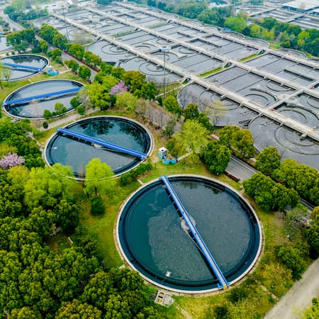 A large wastewater treatment facility with multiple circular and rectangular filtration tanks, surrounded by greenery, representing water purification and environmental sustainability. A large wastewater treatment facility with multiple circular and rectangular filtration tanks, surrounded by greenery, representing water purification and environmental sustainability.